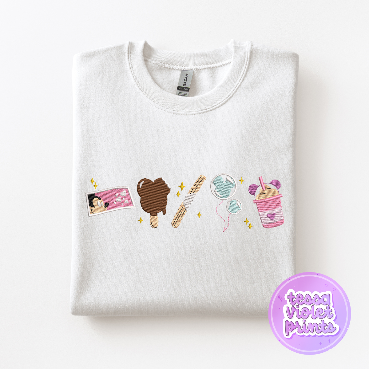 A white t-shirt with a colorful embroidered design depicting various objects, including a brown bear, a pink cup, and a blue balloon.