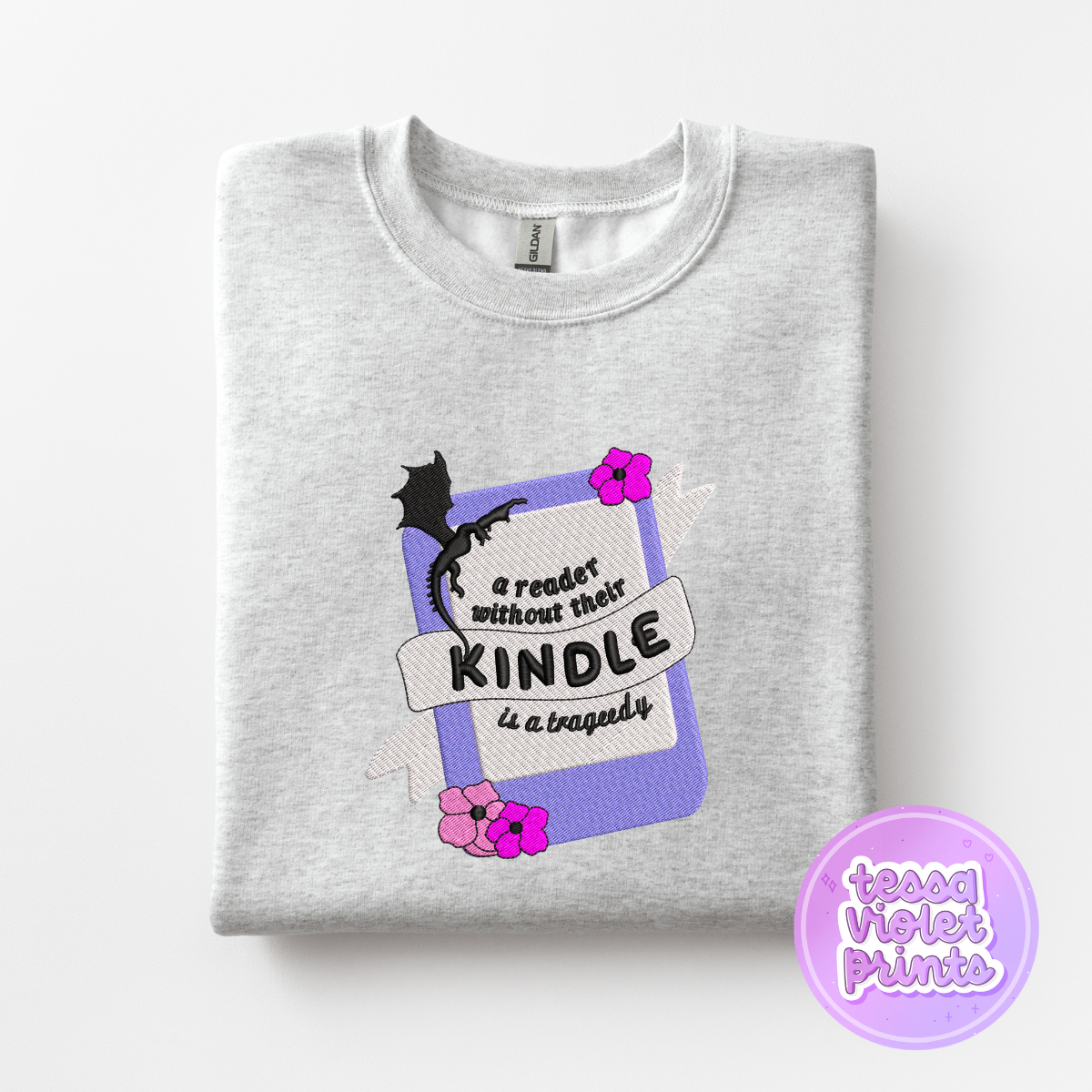 A gray sweatshirt with a graphic design on it, featuring a purple book with a dragon and flowers, along with the text "A reader without their Kindle is a tragedy" and a purple circle with the text "teessa violet prints".