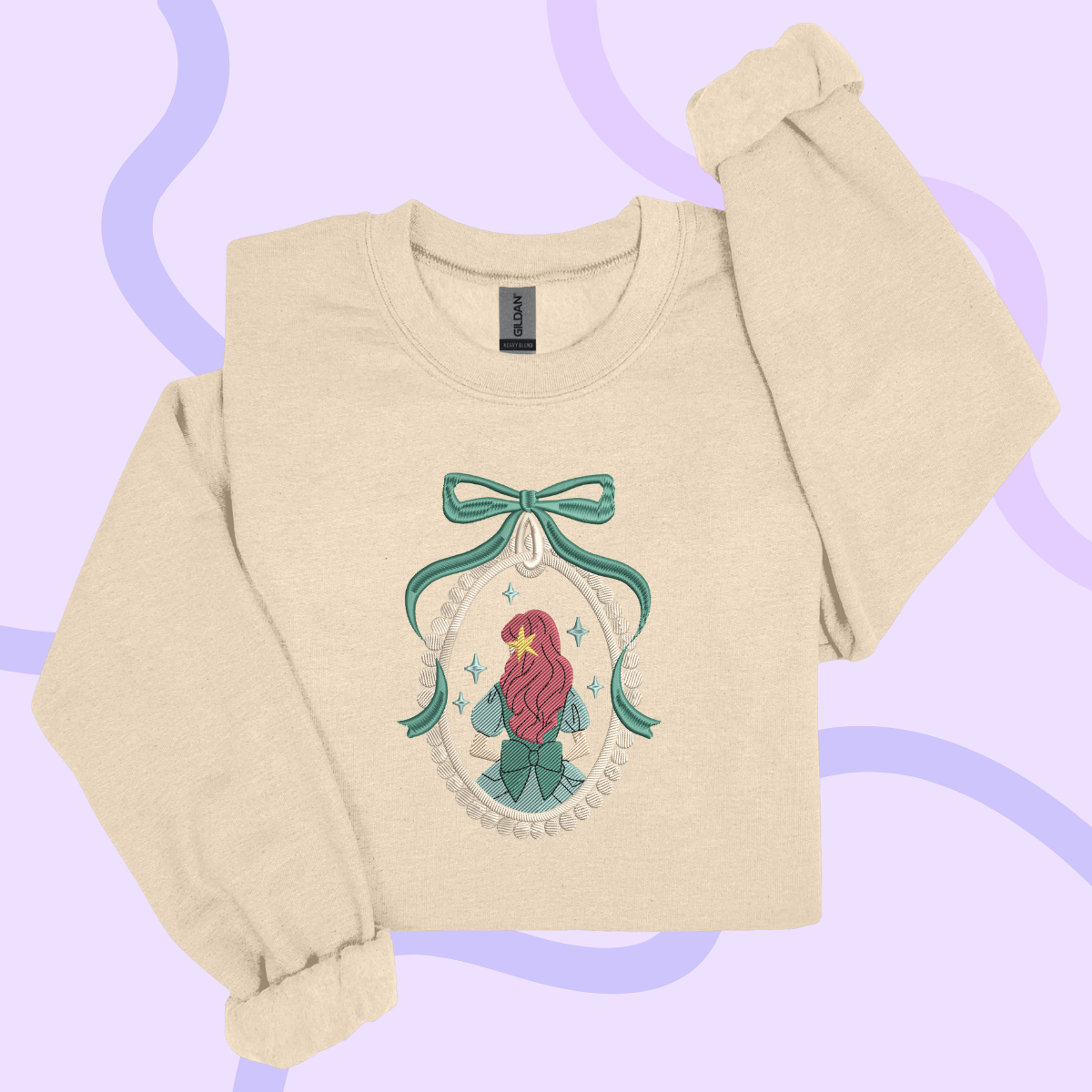 A beige sweater with a graphic design featuring a mermaid or mermaid-like figure, surrounded by a bow and decorative elements, against a purple background.