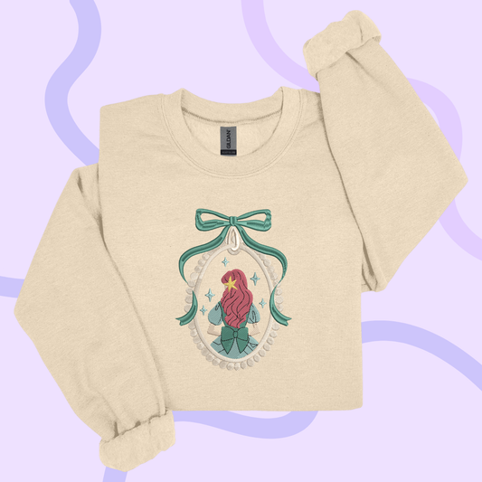 A beige sweater with a graphic design featuring a mermaid or mermaid-like figure, surrounded by a bow and decorative elements, against a purple background.