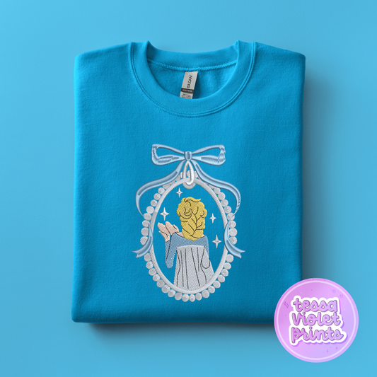 A blue t-shirt with a white embroidered design depicting a woman in a mirror frame with a bow.
