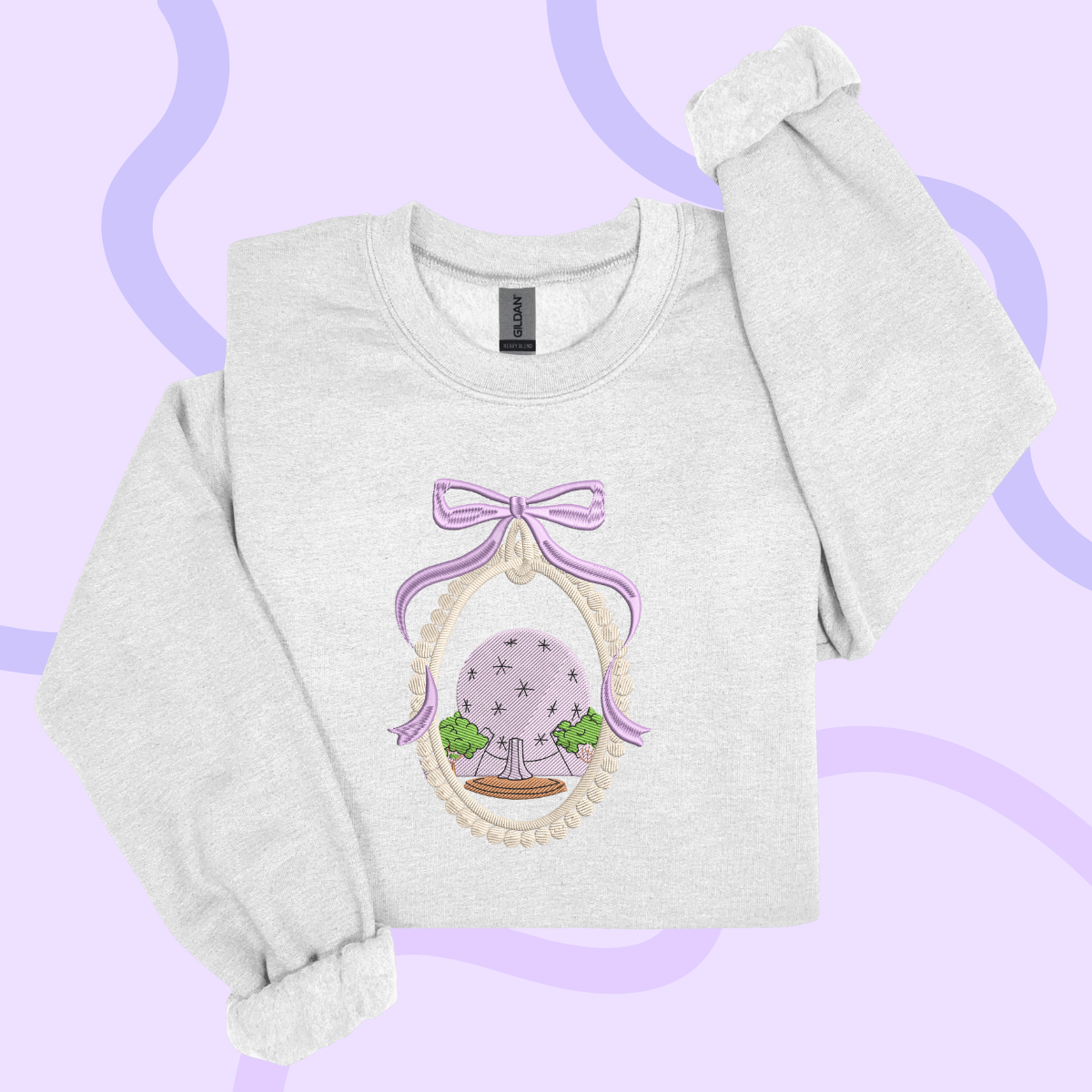 A white sweatshirt with a purple bow and a small, circular embroidered design featuring a starry night sky and a small green plant.