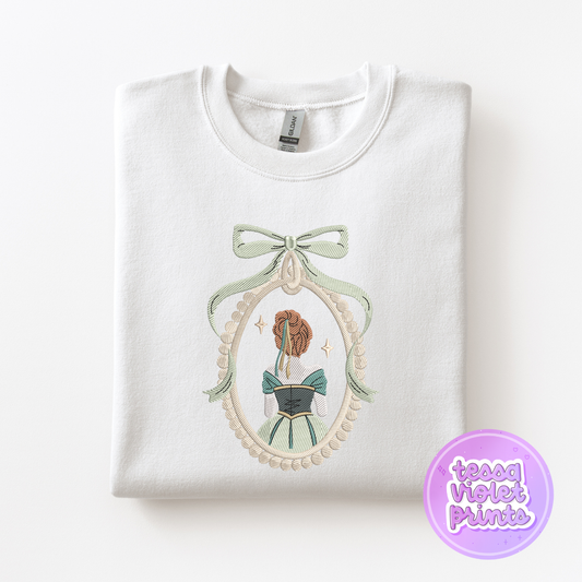 A white sweatshirt with a decorative embroidered design on the front, depicting a woman in a green dress with a bow.