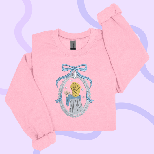 A pink long-sleeved shirt with a blue bow and a small embroidered image of a person wearing a blue dress.