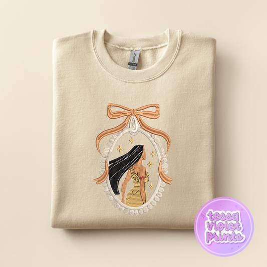 A beige sweatshirt with a decorative embroidered design featuring a woman with long hair and a bow.