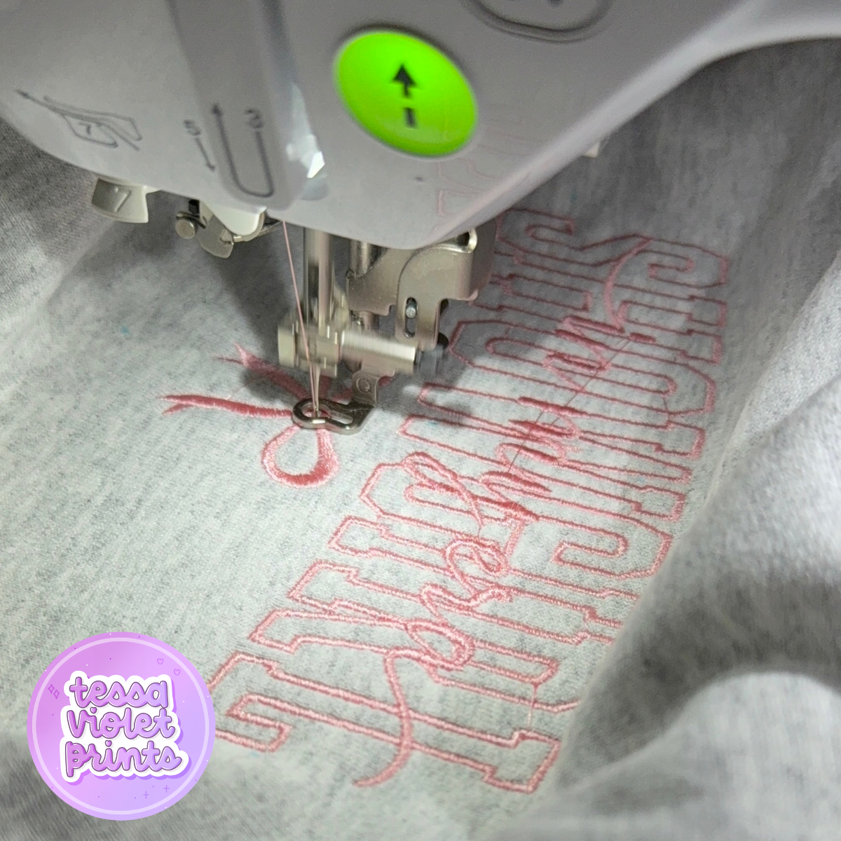 A sewing machine is embroidering a pink design onto a grey fabric.