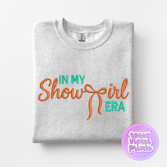 A gray sweatshirt with the text "IN MY SHOW GIRL ERA" printed on it in orange and green colors.