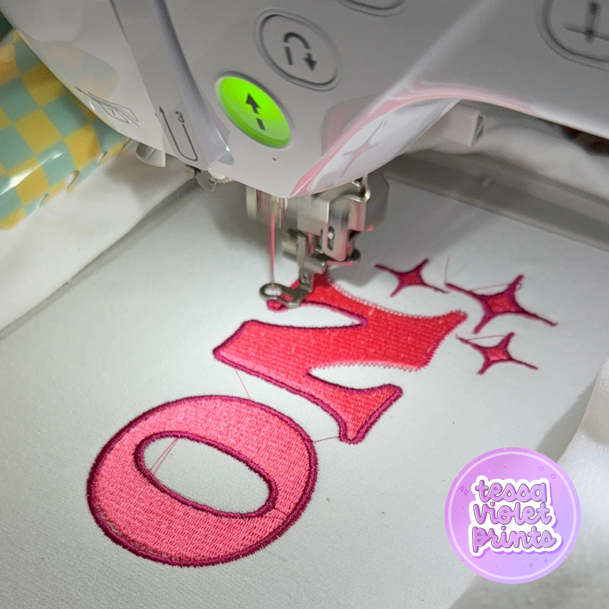 A sewing machine is stitching a large pink letter "O" onto a white fabric, with a starry pattern visible in the background.