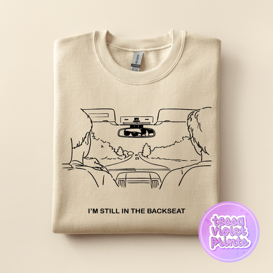 A beige sweatshirt with a black and white line drawing of a car's rearview mirror and two people sitting in the backseat. The text below the drawing reads "I'M STILL IN THE BACKSEAT".