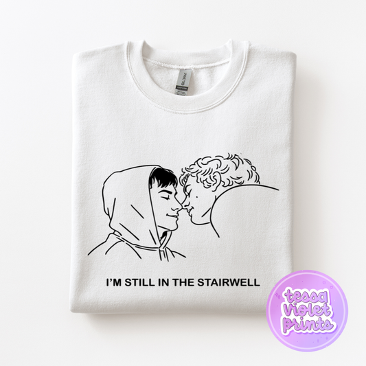 A white t-shirt with a black and white line drawing of two people kissing, accompanied by the text "I'M STILL IN THE STAIRWELL" printed on it.