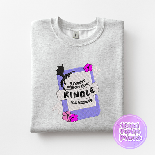 A gray sweatshirt with a graphic design on it, featuring a purple book with a dragon and flowers, along with the text "A reader without their Kindle is a tragedy" and a purple circle with the text "teessa violet prints".