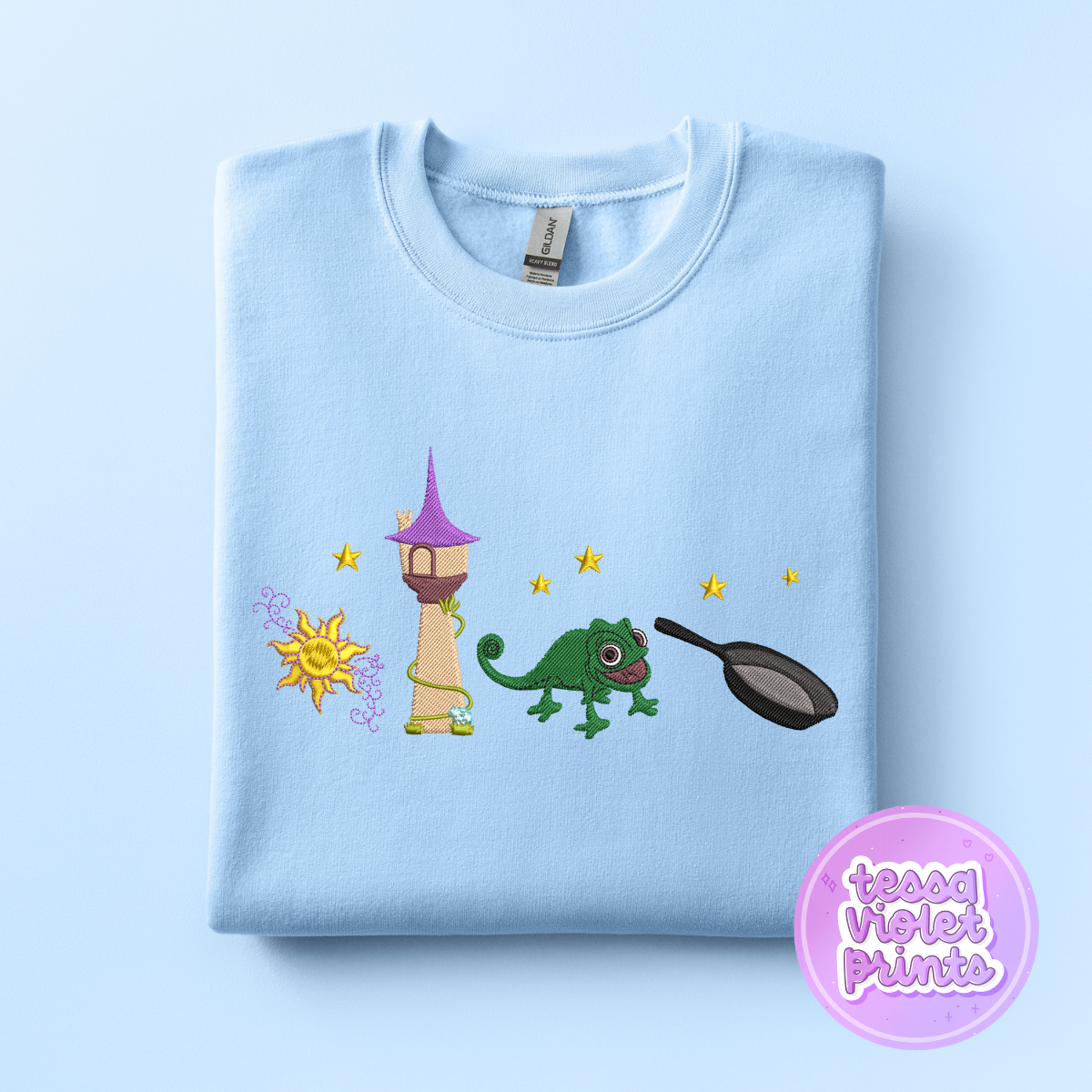 A light blue t-shirt with a colorful embroidered design depicting a castle, a green chameleon, and a frying pan.