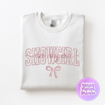A white sweatshirt with the text "SHOWGIRL" printed on it, along with a pink bow.